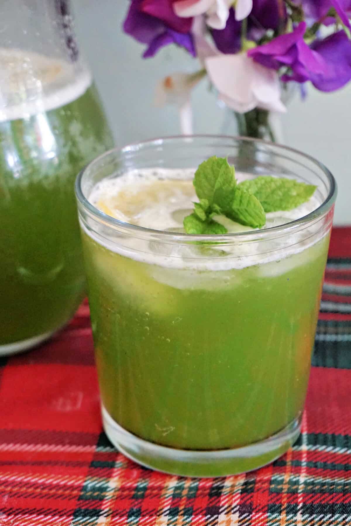 A glass with mint lemonade with partially-visible jug of lemonade and a small vase of flowers in the background.