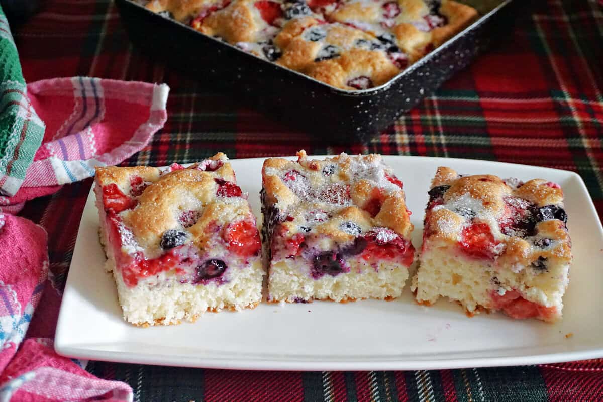 A white rectangle plate with 3 slices of berry cake, and a partially visible tray with cake at the top.
