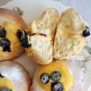 A white plate with 2 halves of a custard and blueberry bun and 3 other partially-visible buns.