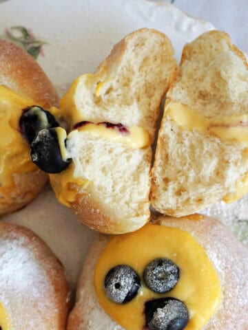 A white plate with 2 halves of a custard and blueberry bun and 3 other partially-visible buns.