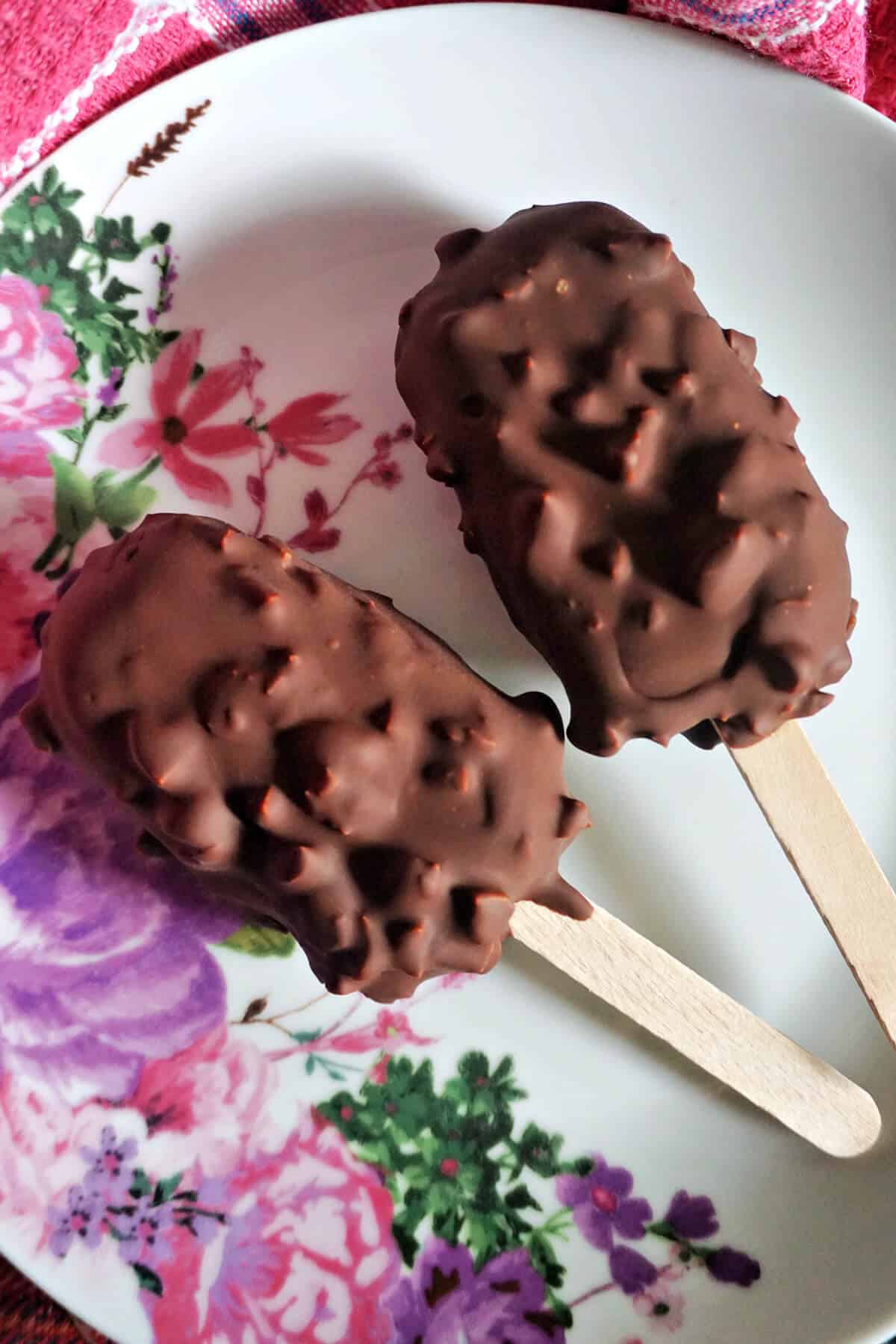 A white flowery plate with 2 chocolate-covered popsicles.