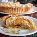A slice of peach tart on a small white flowery plate with a large white plate with the rest of the tart in the background.