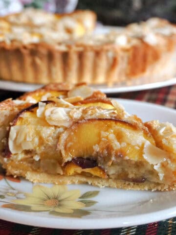 A slice of peach tart on a small white flowery plate with a large white plate with the rest of the tart in the background.