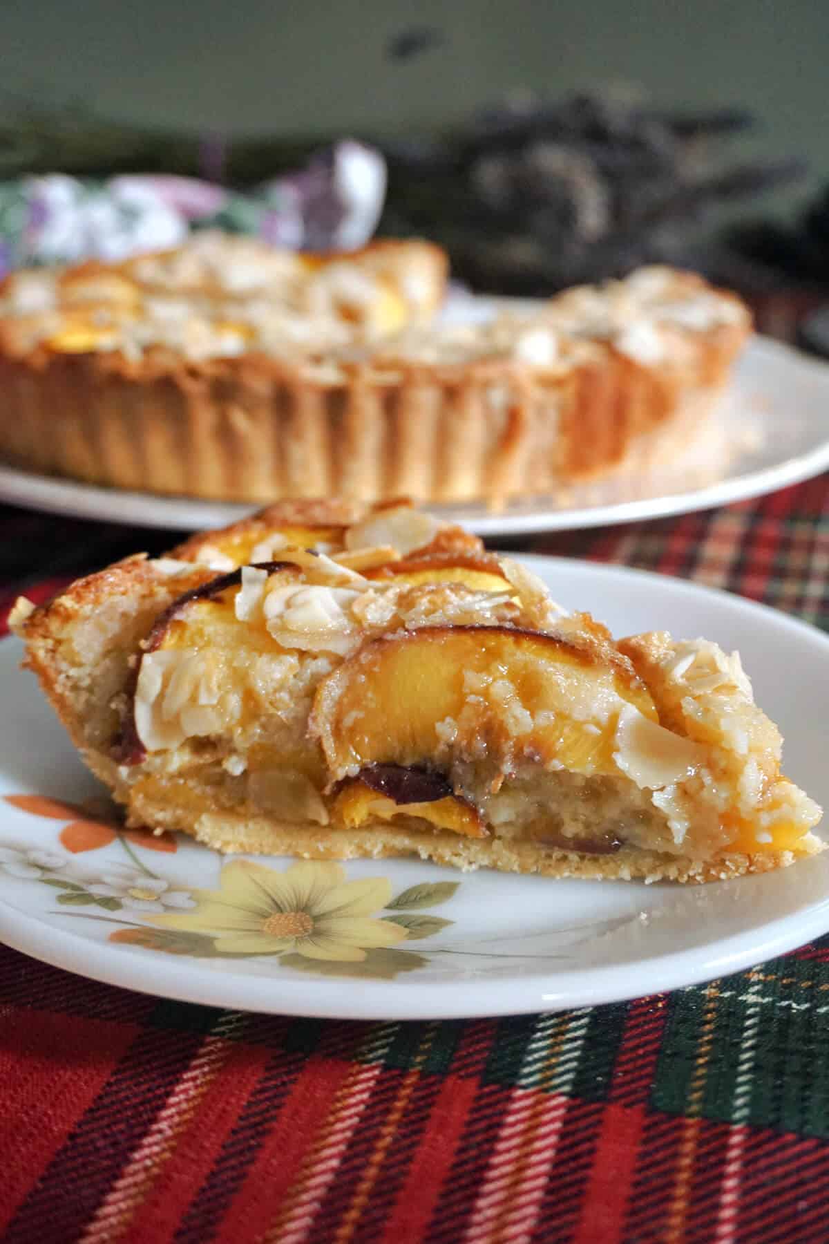 A slice of peach tart on a white dessert plate with a large white plate with the rest of the tart in the background.