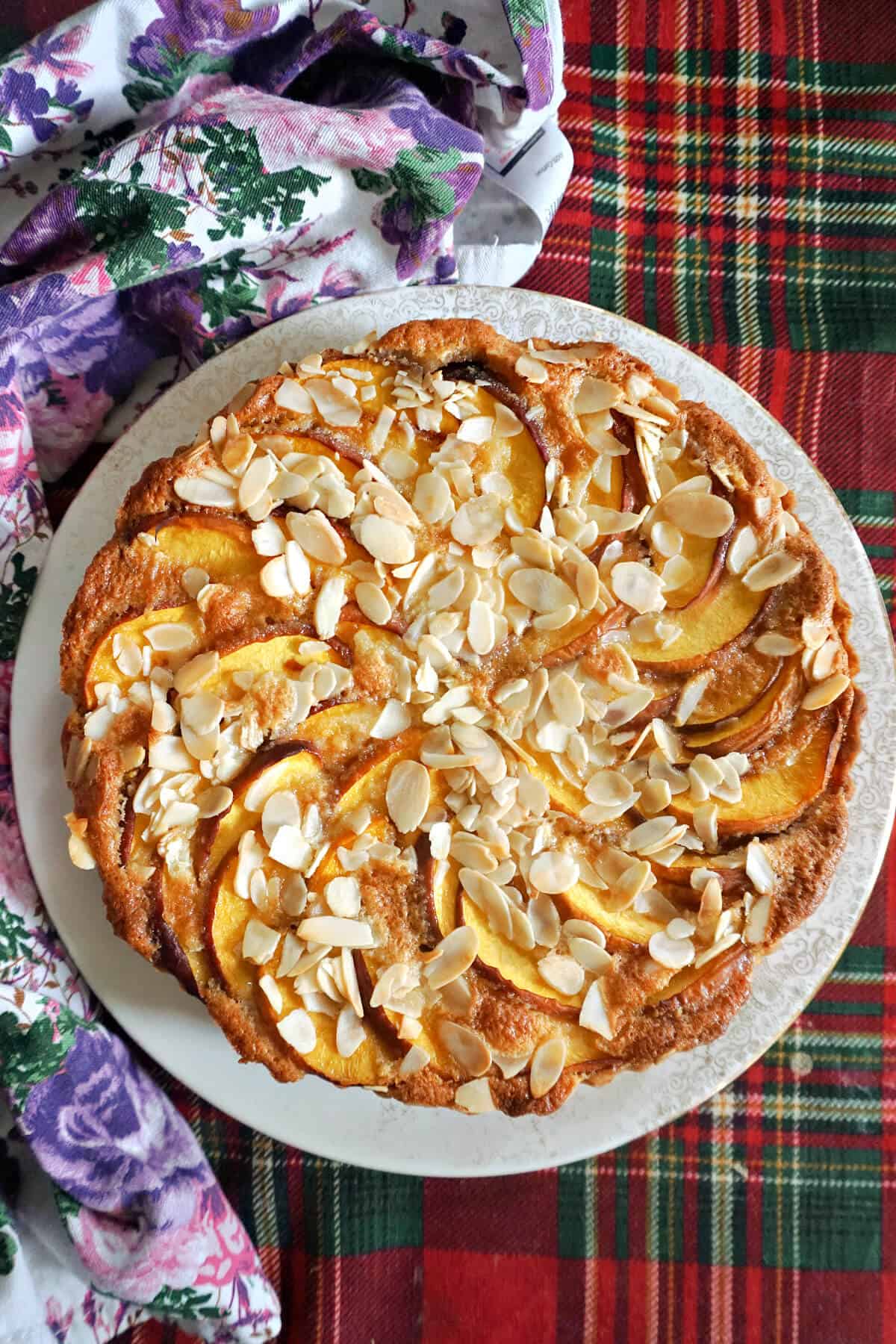 A white plate with a peach almond tart and a flowery kitchen towel around it.