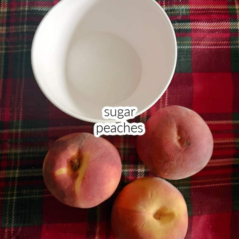 3 fresh peaches and a white small bowl with sugar on a tabletop lined with patterned cloth.