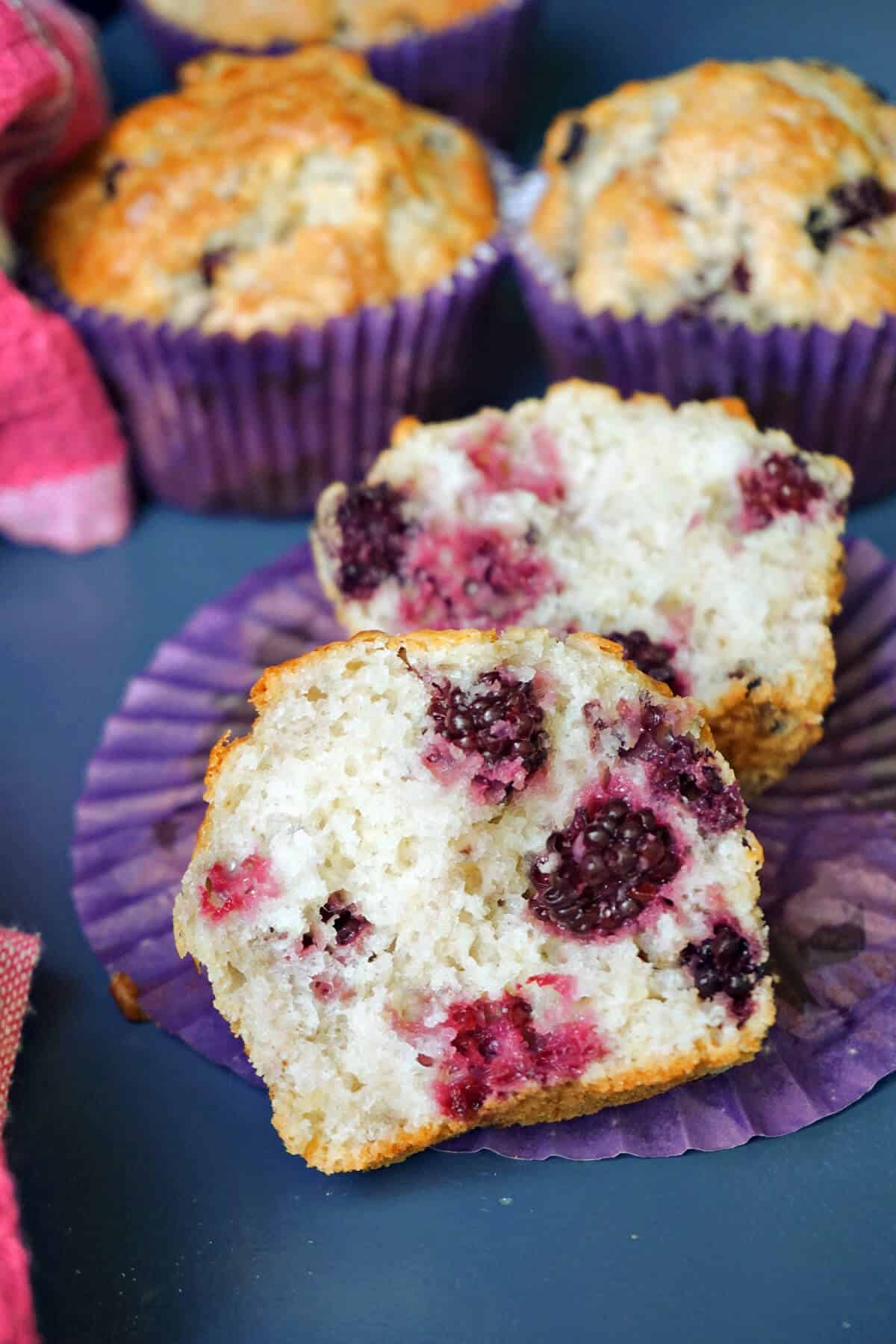 2 halves of a blackberry muffin with other whole muffins partially-visible in the background.
