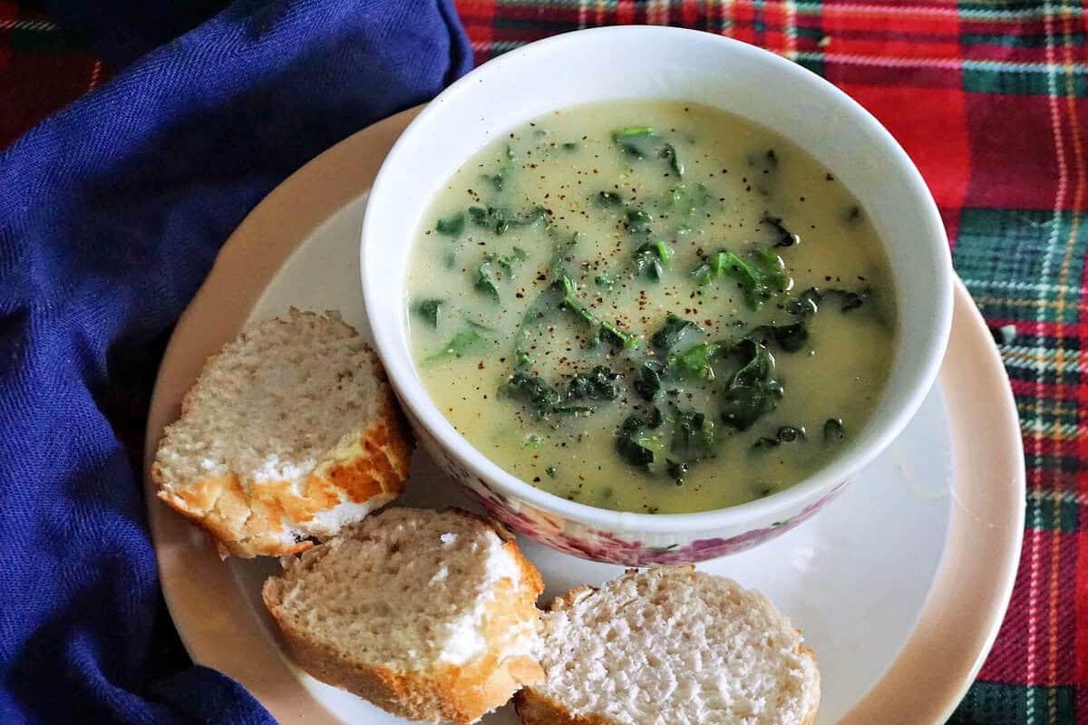 A white bowl with green soup on a white plate with 3 slices of baguette.