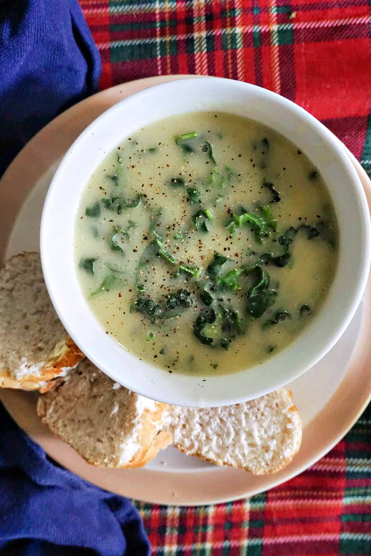 A white bowl with creamy potato and greens soup on a plate with 3 slices of baguette.