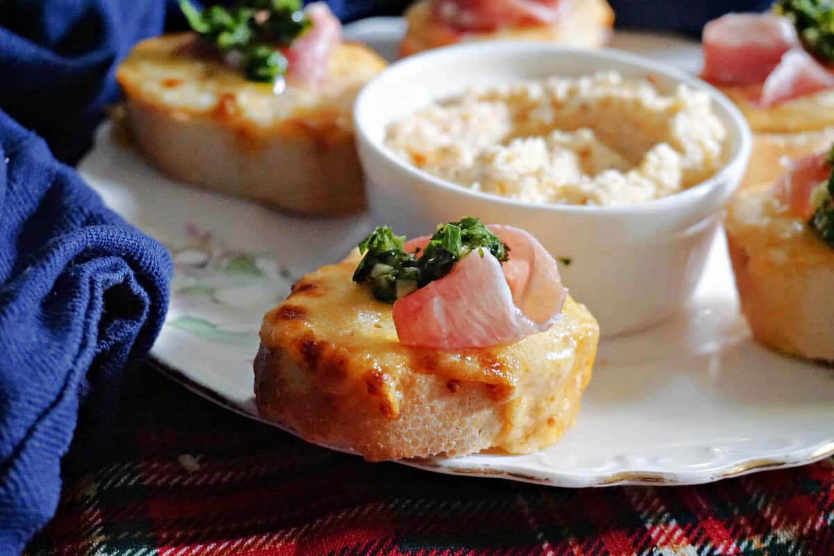 A baguette slice with melted cheese, persillade and prosciutto. In the background a ramekin with cheese spread and other air fried slices.