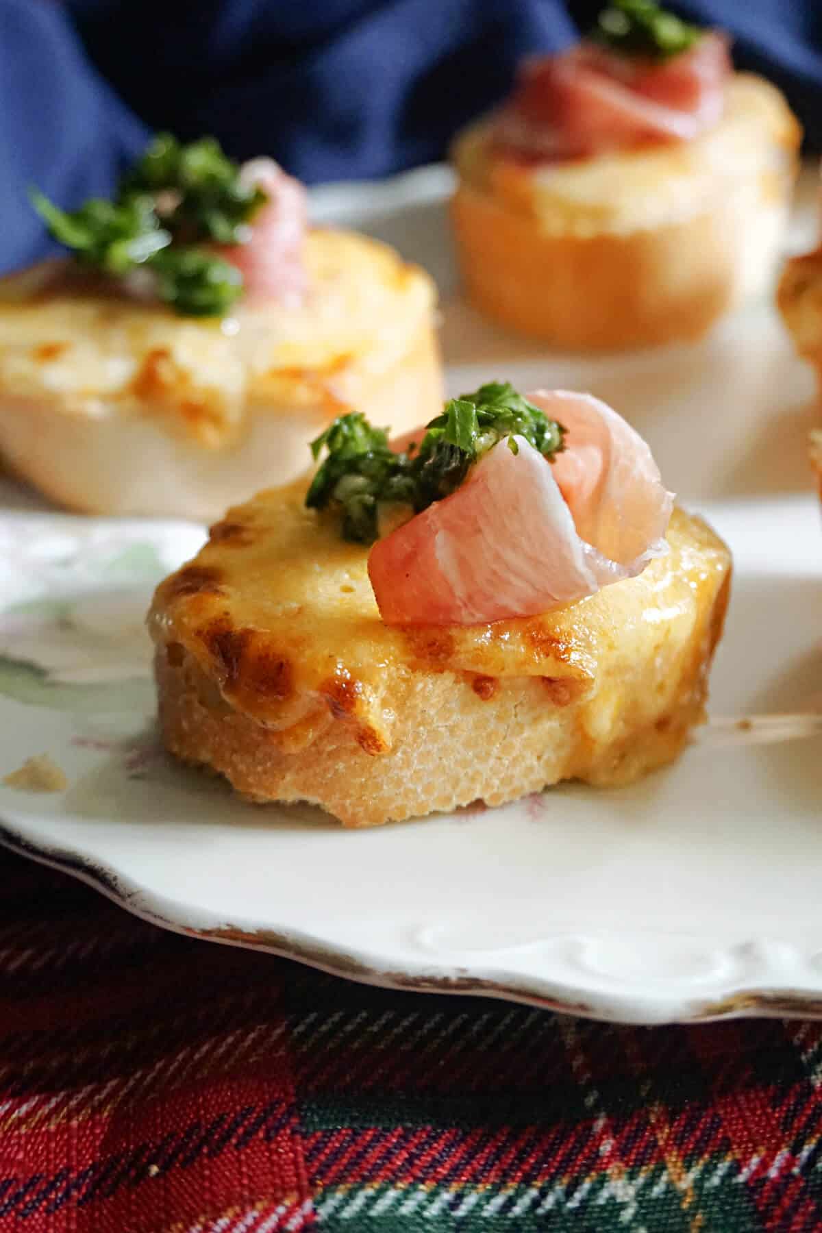 A plate with a slice of baguette topped with melted cheese, prosciutto and parsley sauce and other slices in the background.