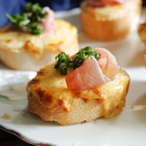 A slice of baguette topped with melted cheese, prosciutto and parsillade.