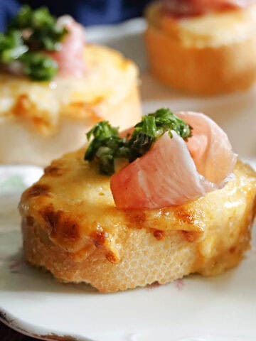 A slice of baguette topped with melted cheese, prosciutto and parsillade.