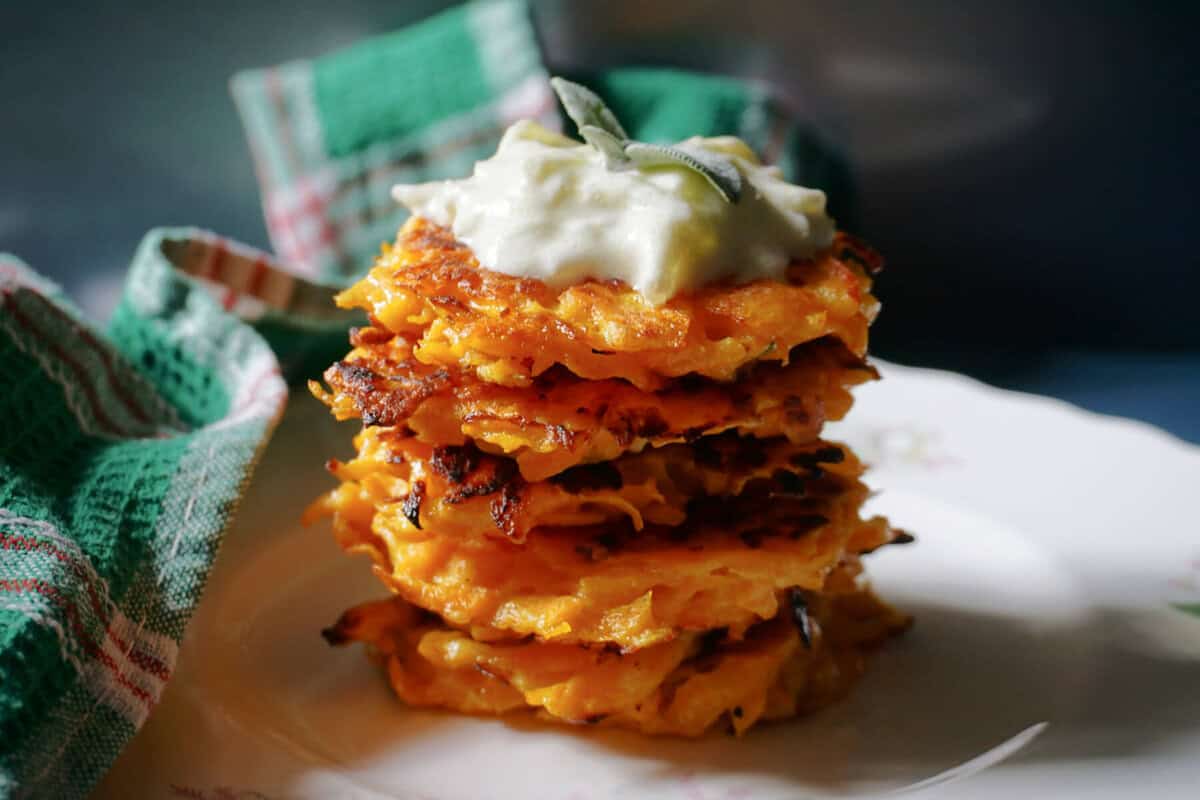 A white plate with a pile of fritters topped with tzatziki and fresh sage leaves. In the background a green patterned tea towel.