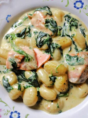 A white plate with a portion of creamy gnocchi with salmon chunks and spinach.
