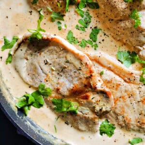 Close-up of a pan with pork steaks smothered in a creamy sauce and garnished with chopped parsley.