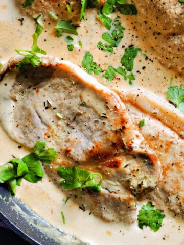 Close-up of a pan with pork steaks smothered in a creamy sauce and garnished with chopped parsley.