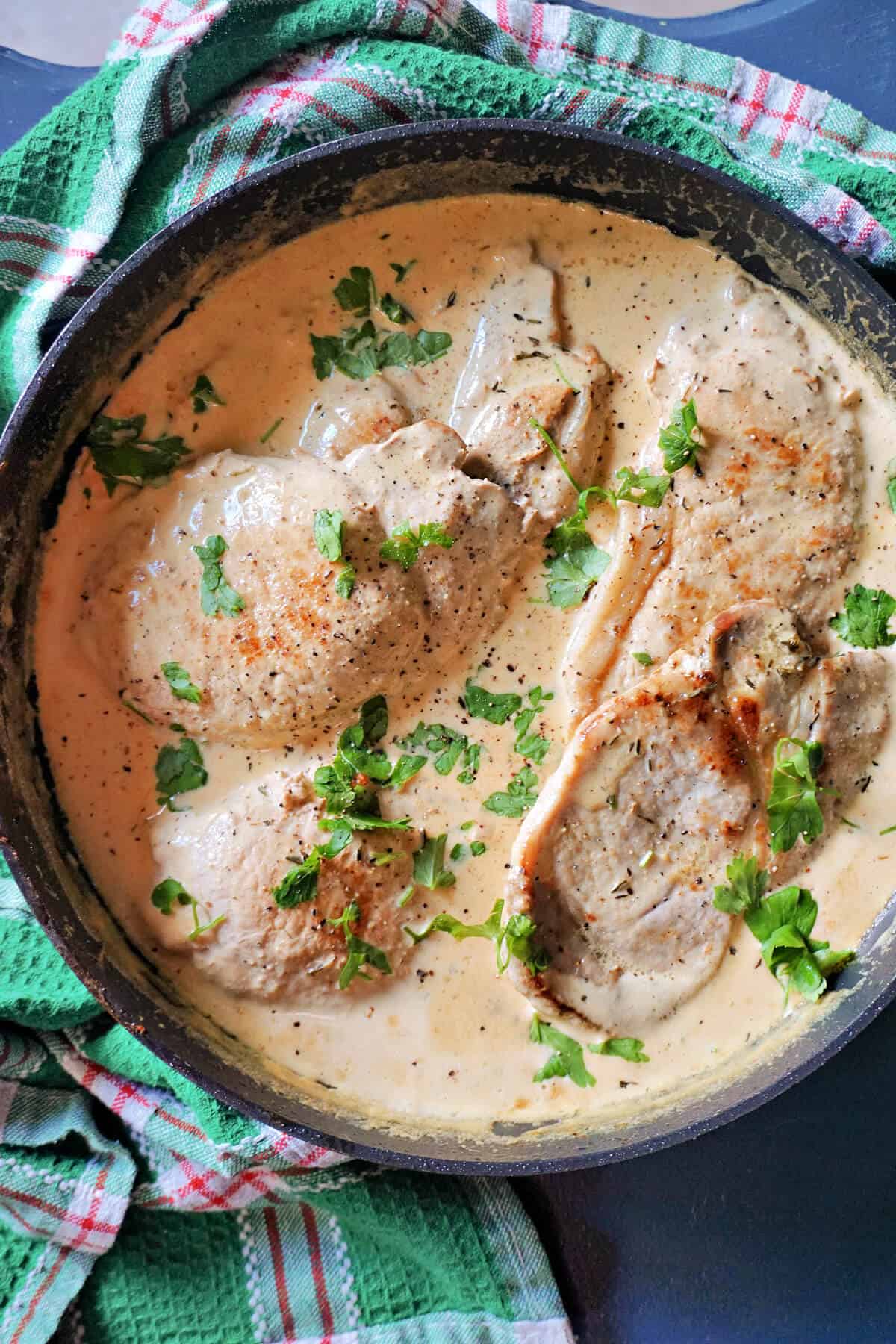 A pan with pork steaks in a creamy sauce, garnished with chopped parsley.